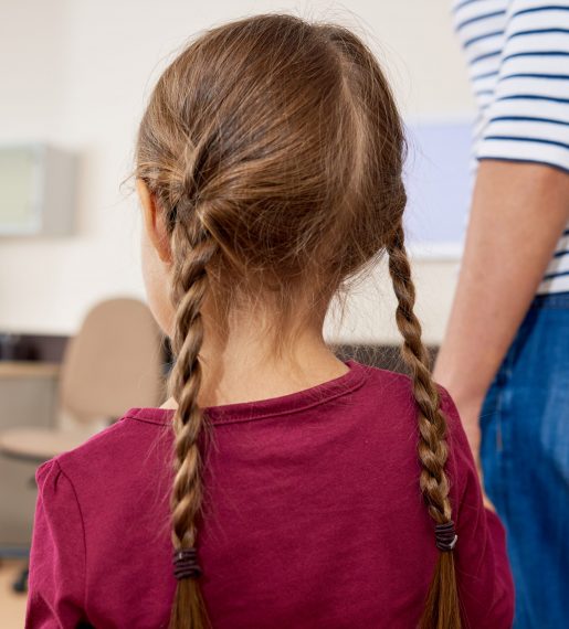 Back view portrait of scared little girl entering doctors office, standing in doorway and holding mothers arm